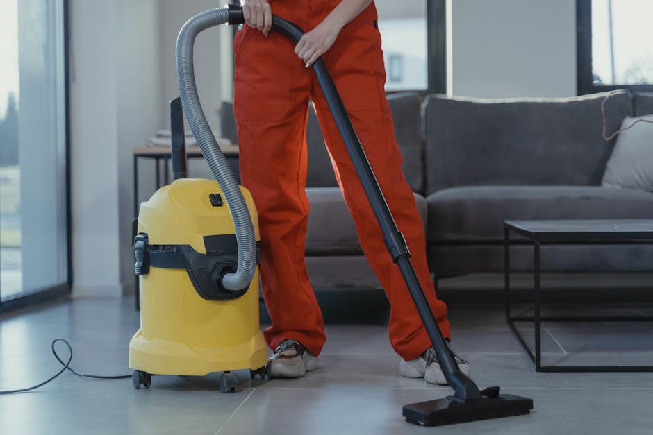 A professional cleaning technician from Carpet Cleaners Bayswater is performing deep cleaning on a beige carpet in a modern living room. The technician is wearing a full white protective suit, including a hood, mask, and gloves, and is using a carpet steam cleaning machine that emits visible steam or mist onto the carpet surface. The living room features a white wall, a large floor mirror leaning against the wall, and a wooden shelving unit with decorative items, plants, and books. The room is well-lit with natural light, emphasizing the cleanliness and freshness of the space, which appears to be in the process of sanitisation or thorough surface cleaning as part of comprehensive domestic cleaning services offered near Queensway Bayswater.