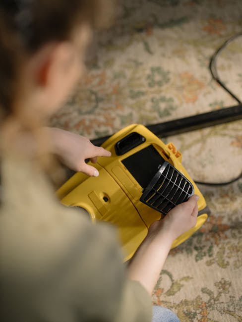 A person holding a yellow portable industrial carpet cleaning machine, focusing on the device with a black ventilation grille and control panel, while working on a patterned area rug in an indoor sett